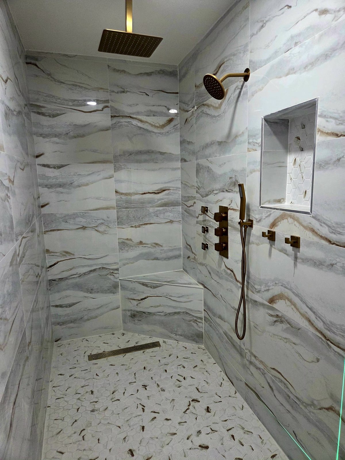 Walk-in shower clad in book-matched marble porcelain with brushed gold rainfall head, body jets and hexagon pebble floor